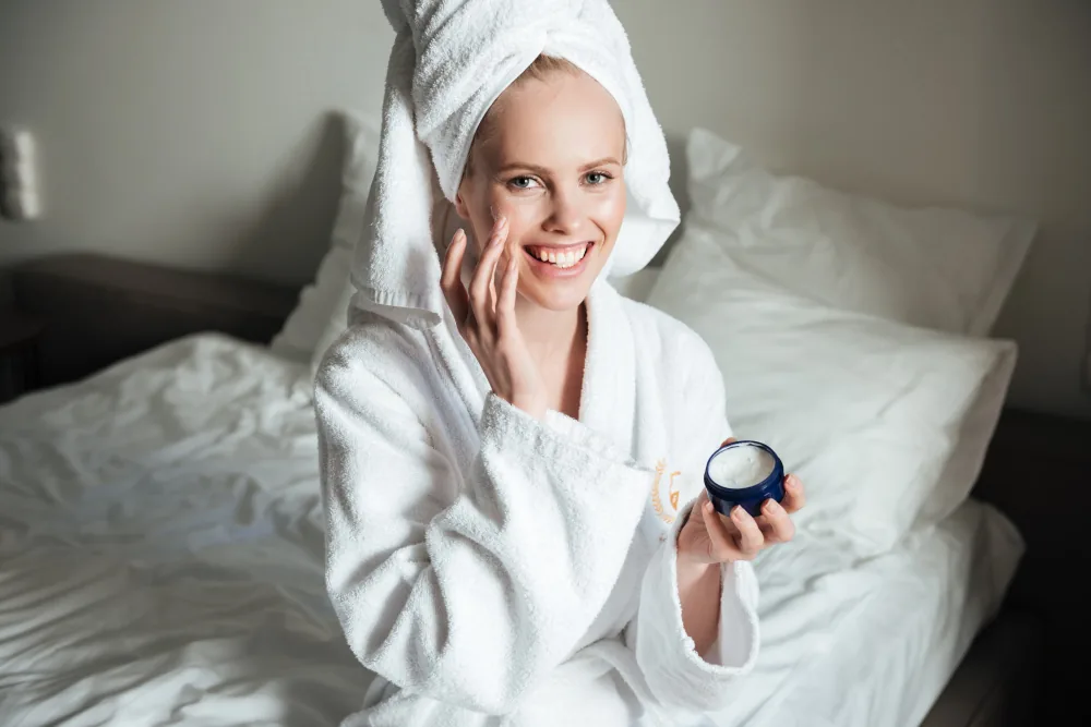 Smiling woman in a bathrobe with a towel wrapped around her hair, sitting on the edge of the bed, holding a jar of face cream in one hand and applying cream to her face with the other