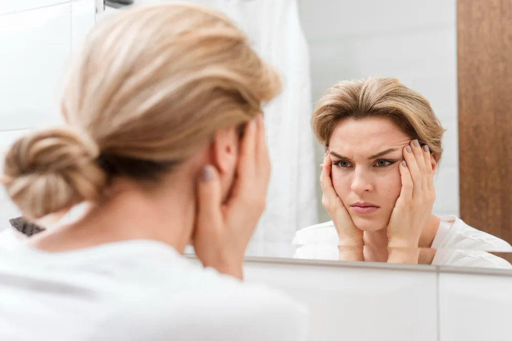 Middle-aged woman in front of a bathroom mirror, raising both hands to her face and anxiously examining her skin