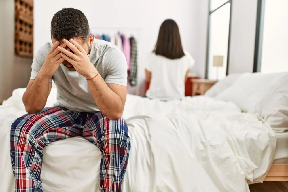 Worried man in pyjamas sitting on the edge of the bed with his face in his hands, his partner sitting on the bed behind him with her back turned