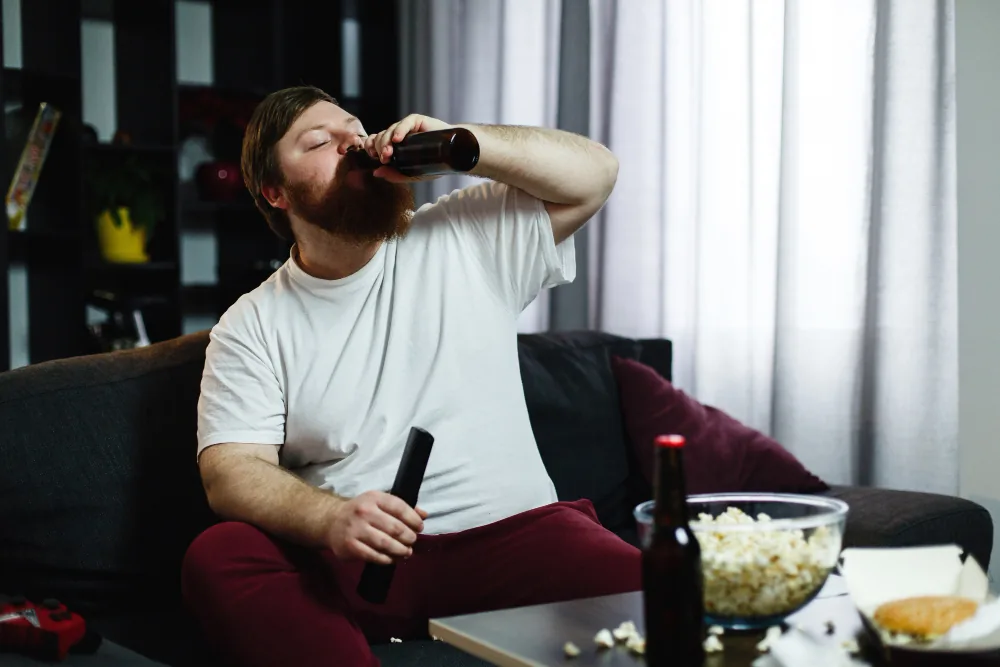Overweight man on a sofa drinking from a beer bottle, with popcorn and a burger in front of him – physical inactivity and excessive drinking are risk factors for a weak erection