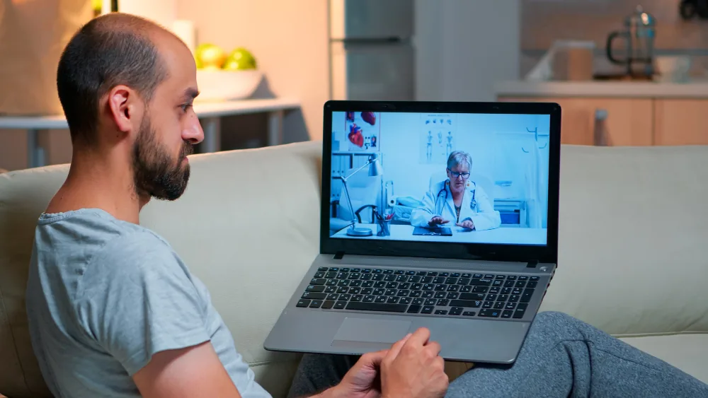 Man sitting on a sofa at home, talking to a female doctor on a laptop