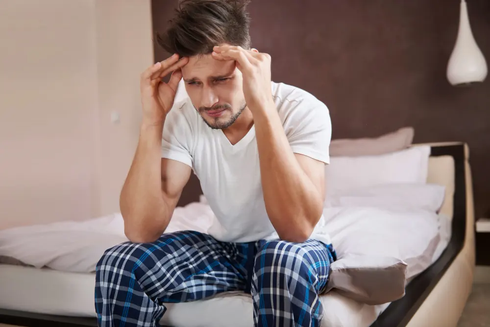 Young man sitting on the edge of a bed, raising his hand to his forehead with a worried expression